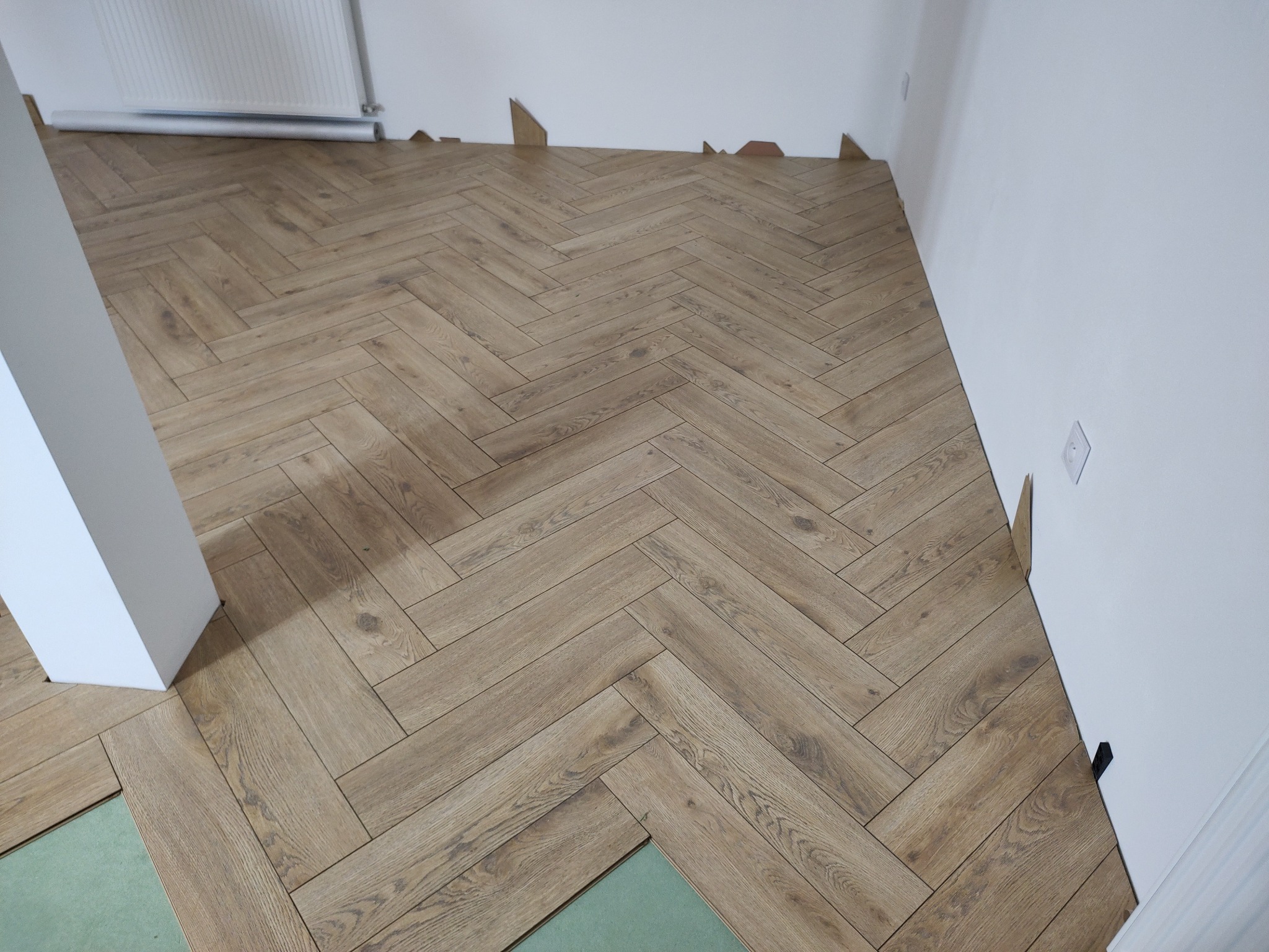 Układanie paneli podłogowych w jodełkę, widoczny fragment podkładu i dylatacja przy ścianie. Jasne panele imitujące drewno, białe ściany i grzejnik.