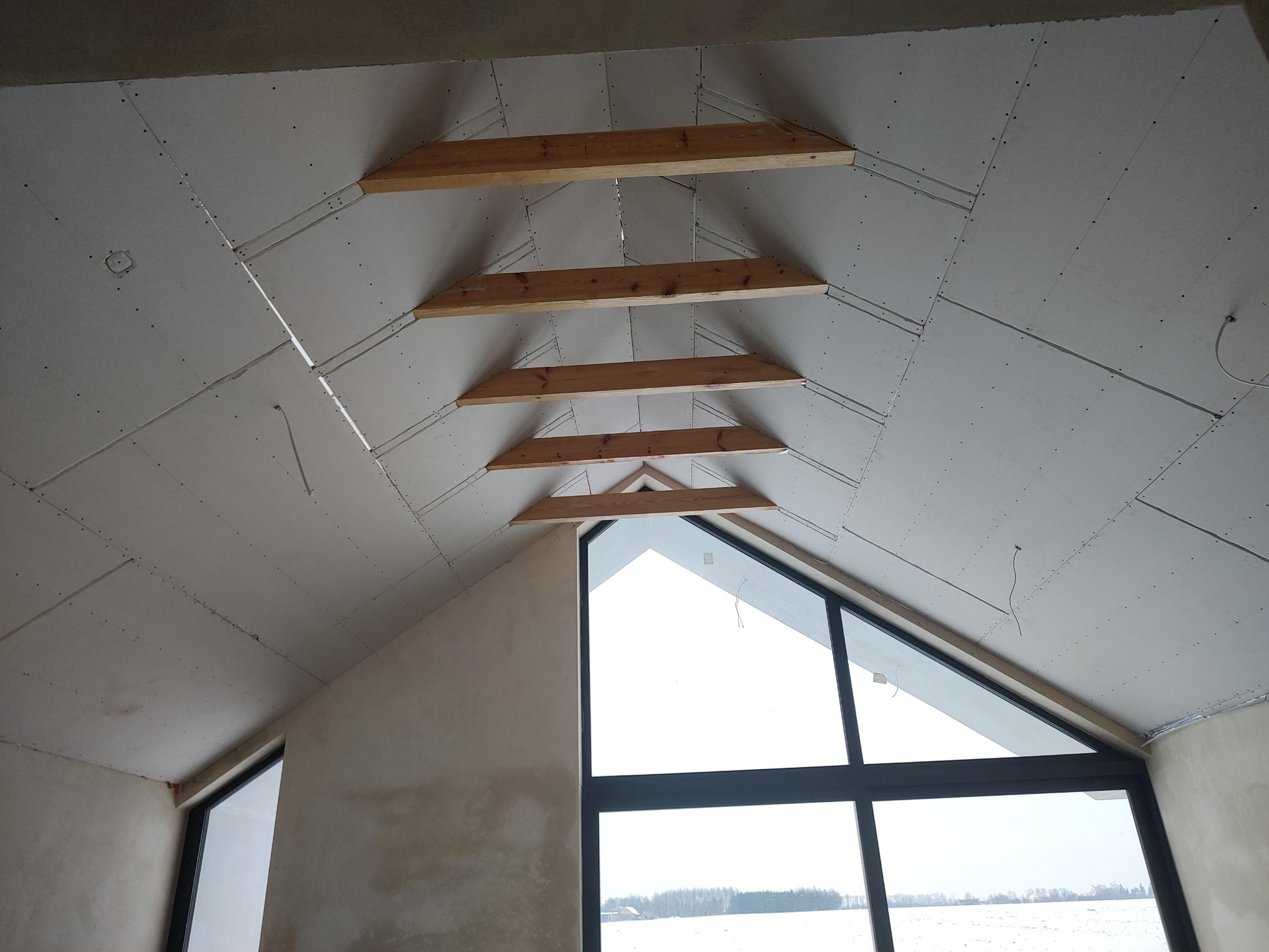 Skośny sufit poddasza z płyt gipsowo-kartonowych, widoczne drewniane belki konstrukcyjne i duże okno z widokiem na zimowy krajobraz. Etap prac wykończeniowych.