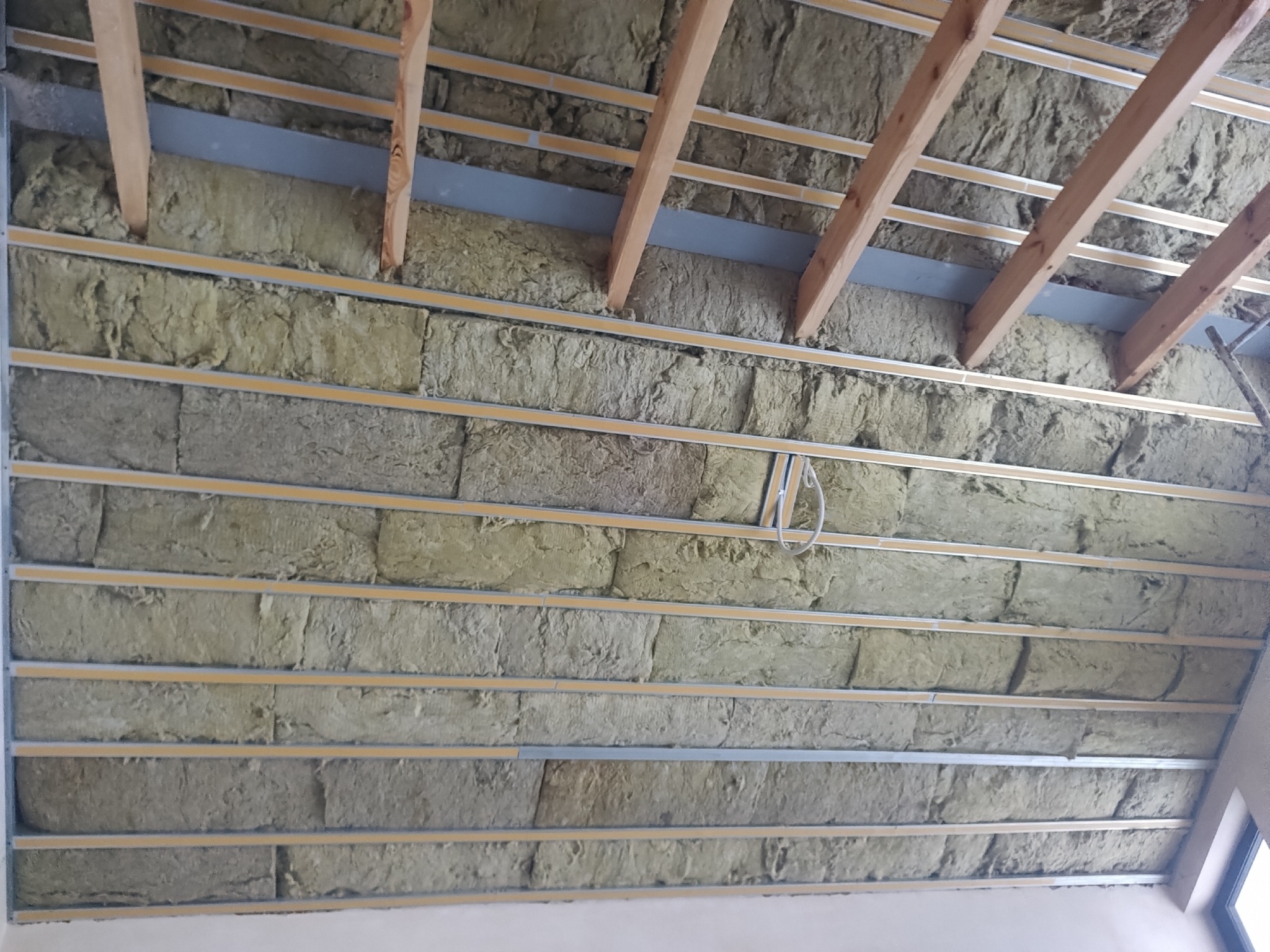 Ocieplenie poddasza wełną mineralną, widoczne drewniane krokwie i metalowe profile. Izolacja termiczna w trakcie montażu, sufit pod skosem.