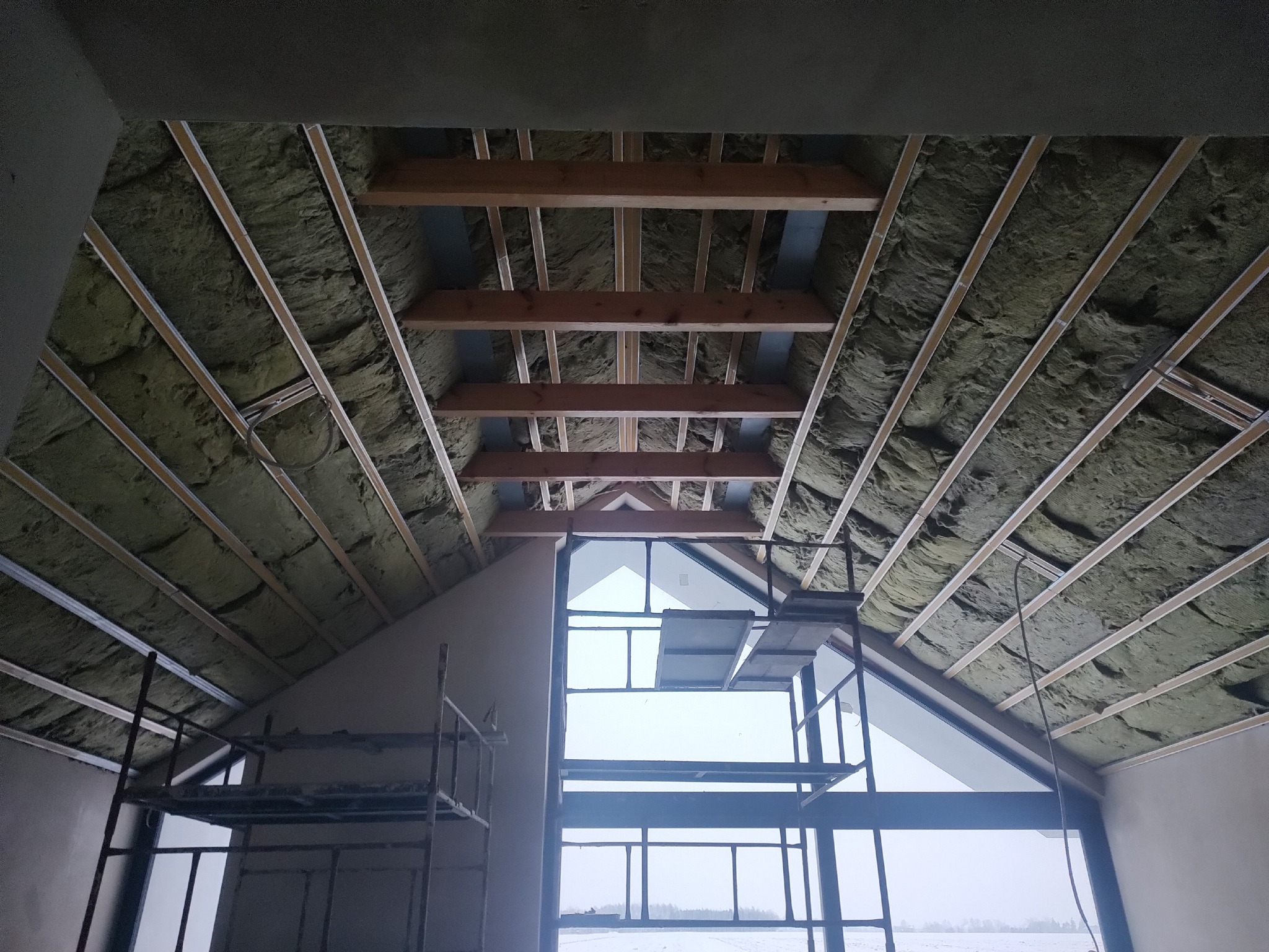 Ocieplenie poddasza wełną mineralną, widoczne drewniane elementy konstrukcyjne i rusztowanie. Prace wykończeniowe w nowym domu z dużym oknem.