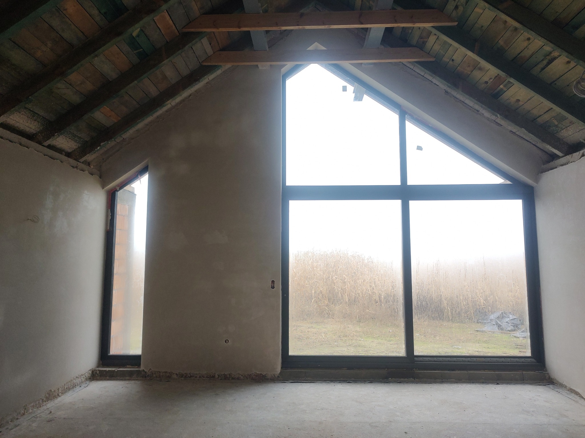 Surowe wnętrze poddasza z oknami, ściany po wstępnym tynkowaniu, widoczne drewniane elementy konstrukcji dachu. Za oknami pole w porannej mgle.