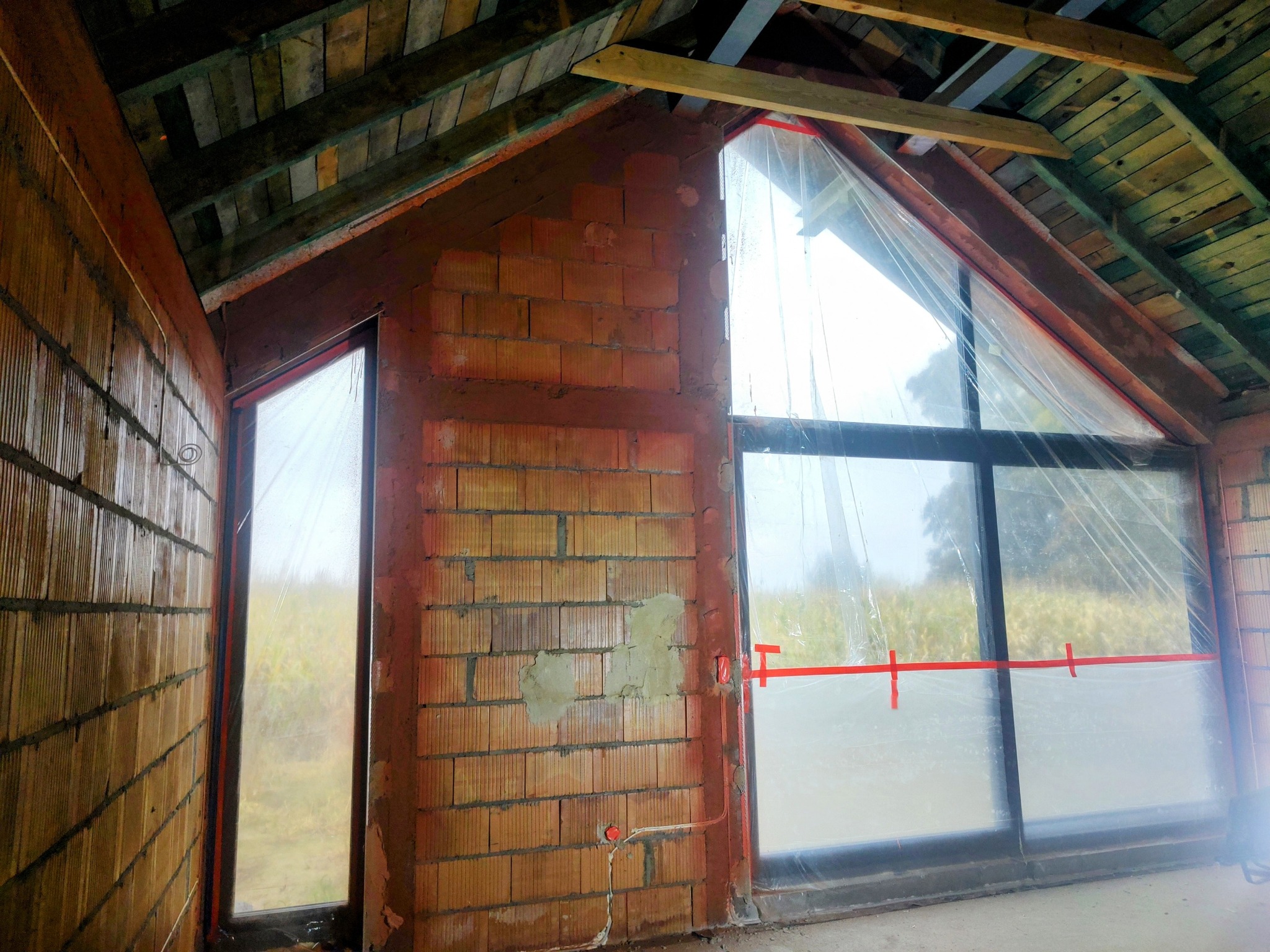 Surowy stan poddasza z ceglanymi ścianami, zabezpieczonymi oknami folią i drewnianą konstrukcją dachu, ukazujący etap przygotowania do dalszych prac wykończeniowych.