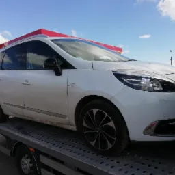 Biały samochód Renault Scenic na platformie lawety, widoczny na tle stacji benzynowej w słoneczny dzień.