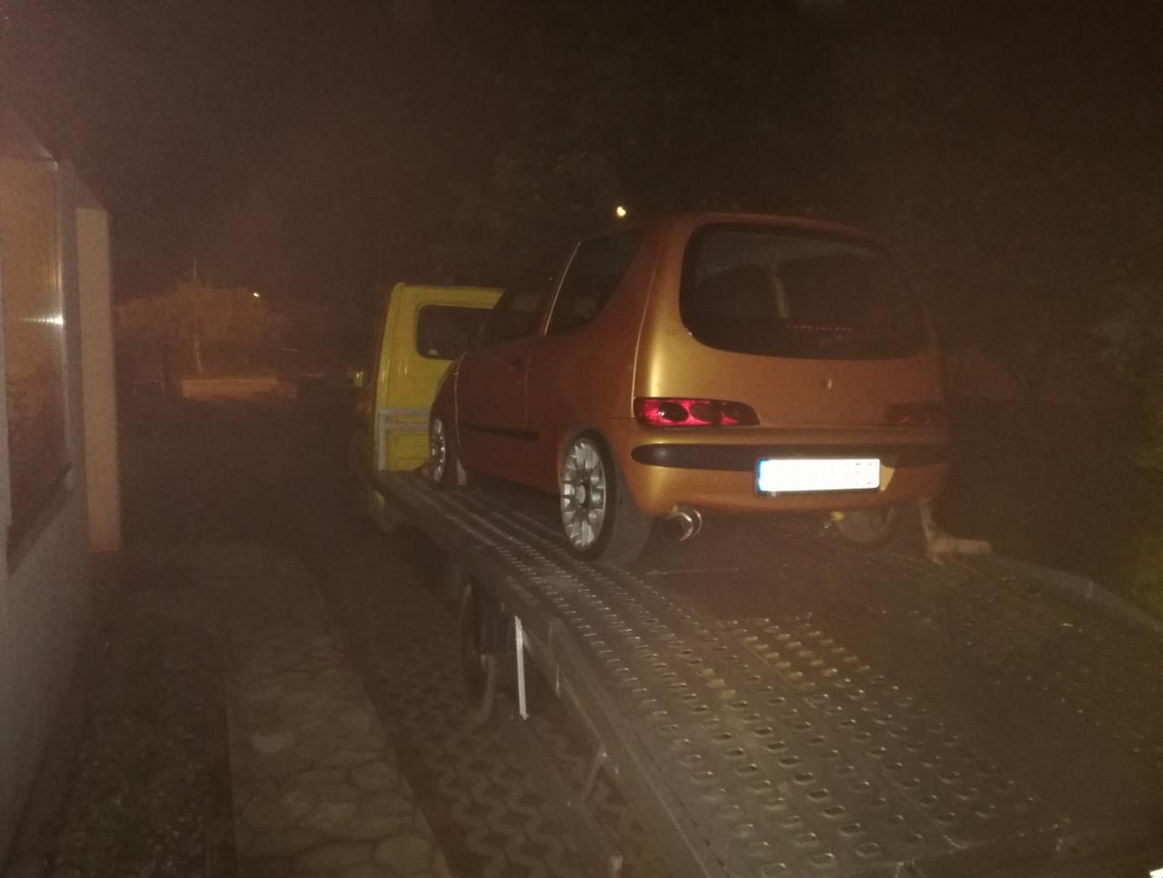 Pomarańczowe auto marki Fiat Seicento z tuningowanym wydechem, zabezpieczone pasami, załadowane na żółtą platformę pojazdu transportowego w słabym oświetleniu nocnym.