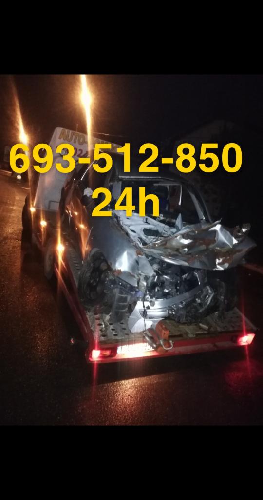 Uszkodzony samochód osobowy, z widocznymi zniszczeniami karoserii, załadowany na platformę pojazdu pomocy drogowej z napisem 'AUTO' i numerem telefonu '693-512-850 24h' w słabych warunkach...