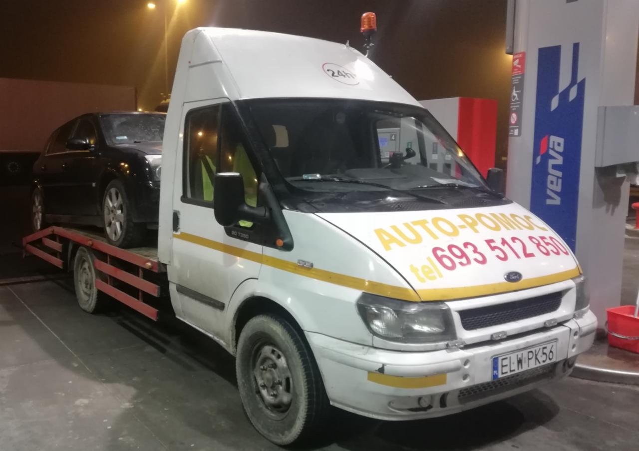 Czarny samochód osobowy na platformie białej autolawety Ford Transit z napisem AUTO-POMOC i numerem telefonu, stojącej na stacji benzynowej. Widoczna tablica rejestracyjna ELW PK56.