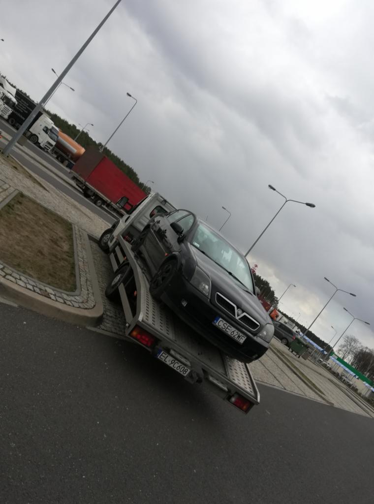 Czarny samochód osobowy z uszkodzonym przodem, zabezpieczony na platformie lawety w Koluszkach, widok z boku na tle pochmurnego nieba i parkingu dla tirów.