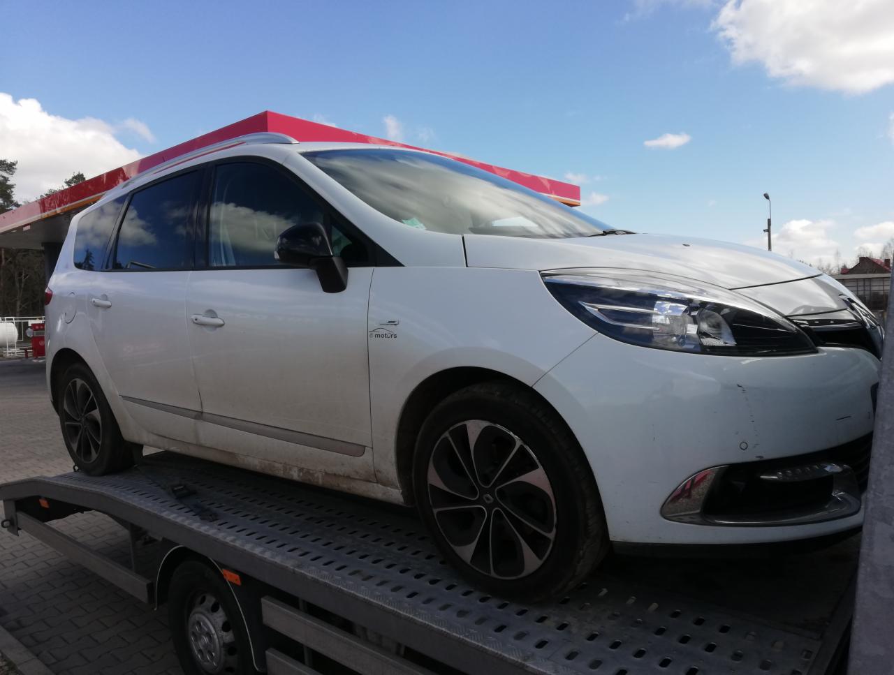Biały samochód Renault Scenic na platformie lawety, widoczny na tle stacji benzynowej w słoneczny dzień.