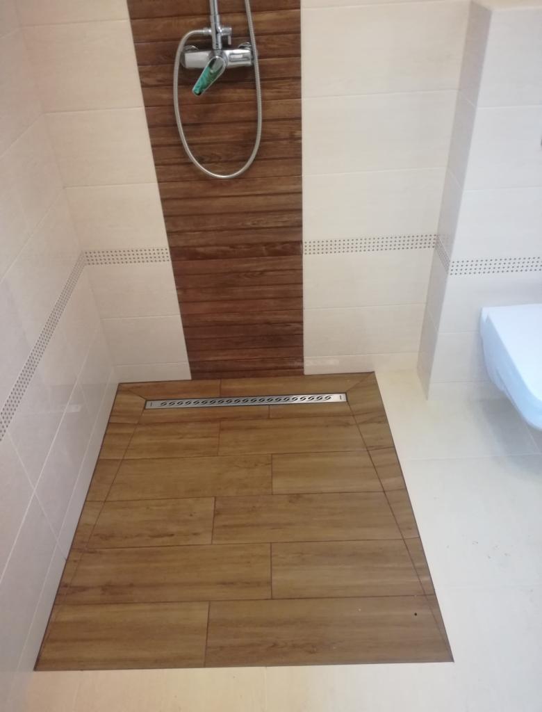 Wykończony brodzik prysznicowy z odpływem liniowym, wyłożony płytkami imitującymi drewno, z baterią natryskową na ścianie z pasem drewnopodobnych paneli.