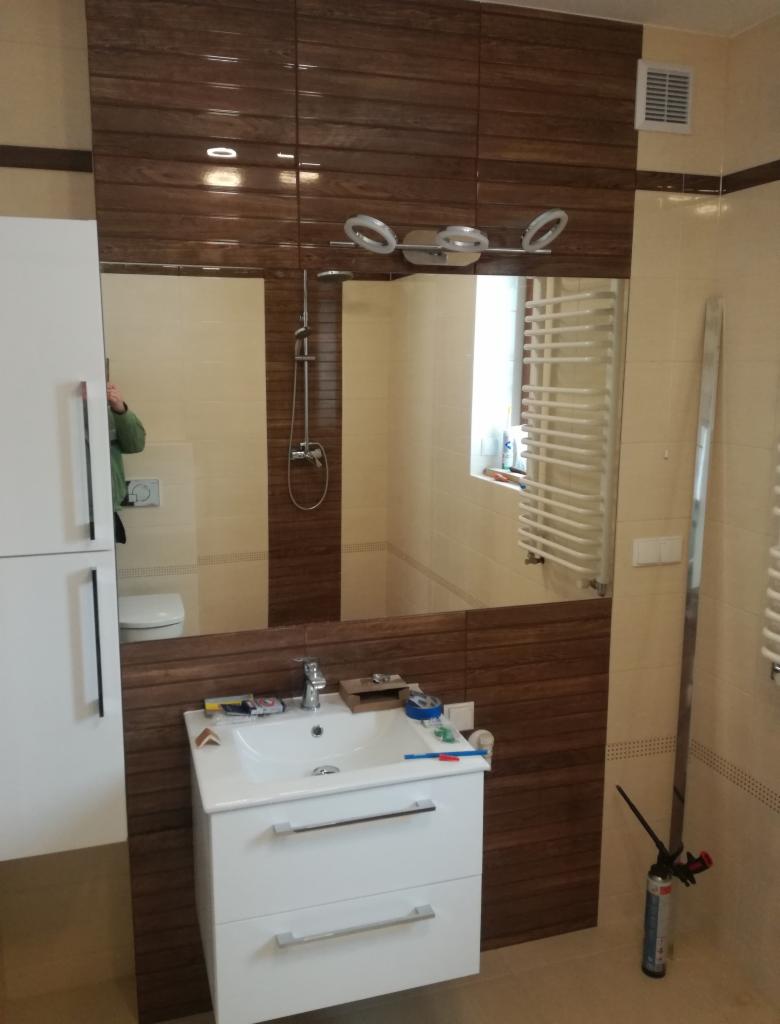 Wykończona łazienka z brązowymi płytkami imitującymi drewno, białą szafką pod umywalką i dużym lustrem, widoczny grzejnik drabinkowy i pistolet do pianki montażowej.