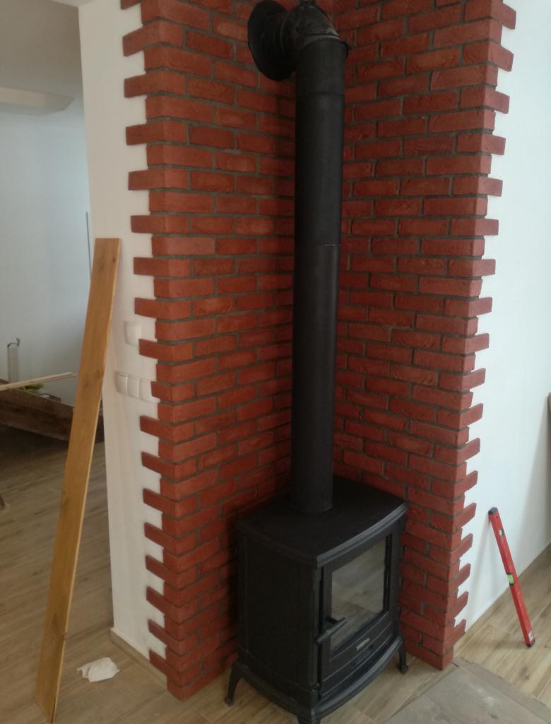 Czarny piec wolnostojący z rurą kominową umieszczony w rogu wykończonym czerwoną cegłą klinkierową, widoczna poziomica i deska.