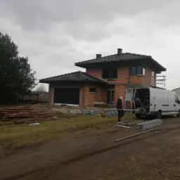 Budynek w trakcie budowy z widocznym dachem pokrytym dachówką, pracownikiem wyładowującym elementy konstrukcyjne z białego furgonetki i stosem drewna na trawniku w pochmurny dzień.