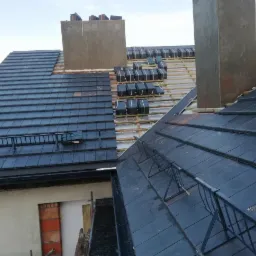 Układanie ciemnej dachówki ceramicznej na dachu z widocznymi kominami w trakcie prac, zabezpieczenia na dachu.