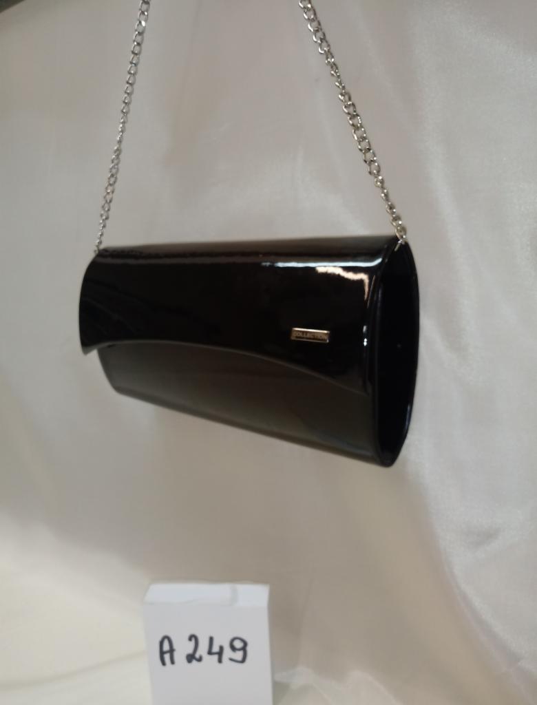 Czarna, lakierowana torebka wizytowa na srebrnym łańcuszku, zawieszona na tle białej, lekko pogniecionej tkaniny. Na pierwszym planie widoczna kartka z napisem 'A 249'.