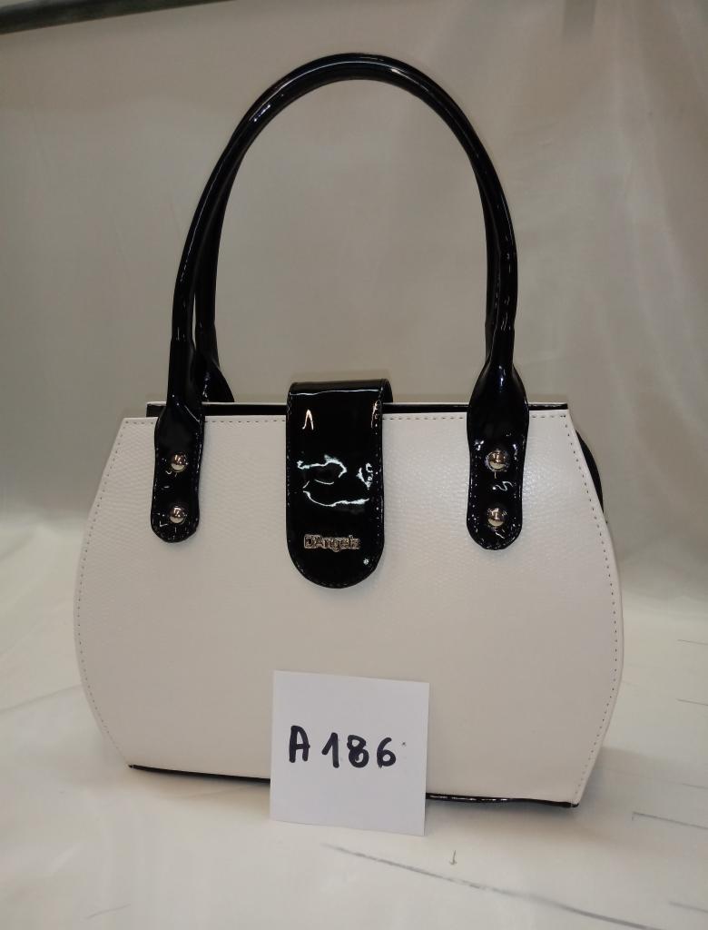 Biała torebka damska z czarnymi, lakierowanymi uchwytami i klapką, z widoczną kartką z numerem 'A 186', na jasnym tle.