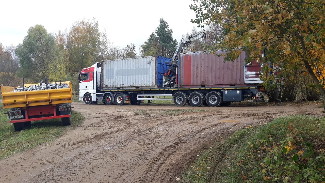 Ciężarówka z dwoma kontenerami na naczepie i dźwigiem, obok żółta przyczepa wypełniona metalowymi rurami, na błotnistej drodze.