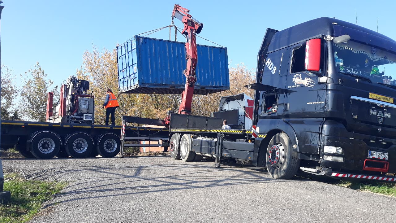 Ciężarówka z dźwigiem przenosi niebieski kontener; pracownik w pomarańczowej kamizelce stoi na platformie, kontrolując operację.