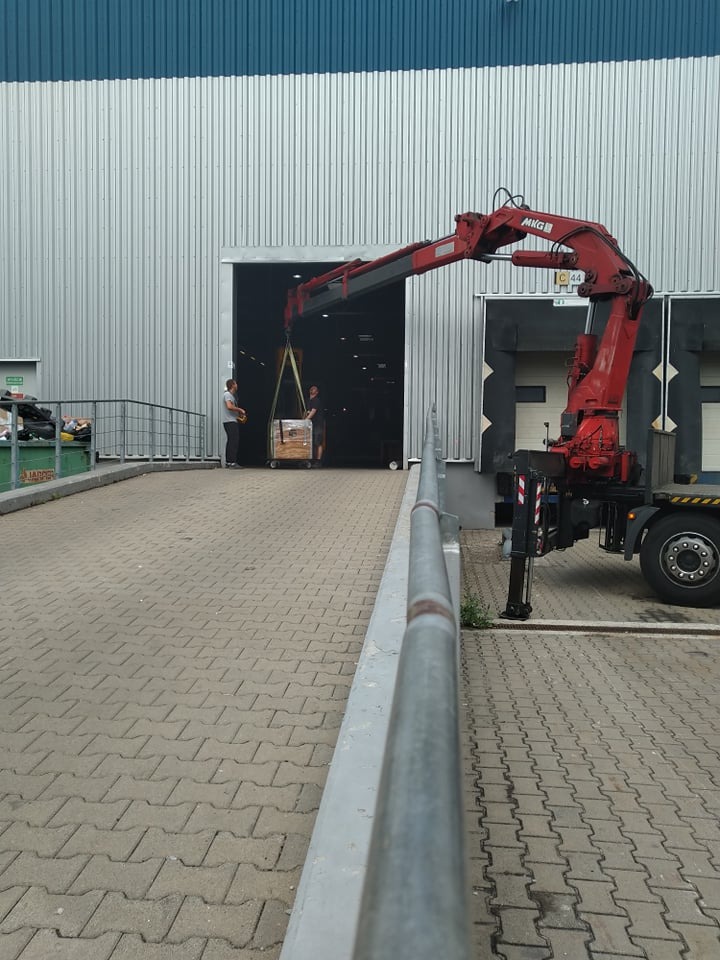 Czerwony dźwig samochodowy MKG podnosi paletę z towarem przed magazynem z dwoma pracownikami w tle, widoczne kostki brukowe i metalowa barierka.