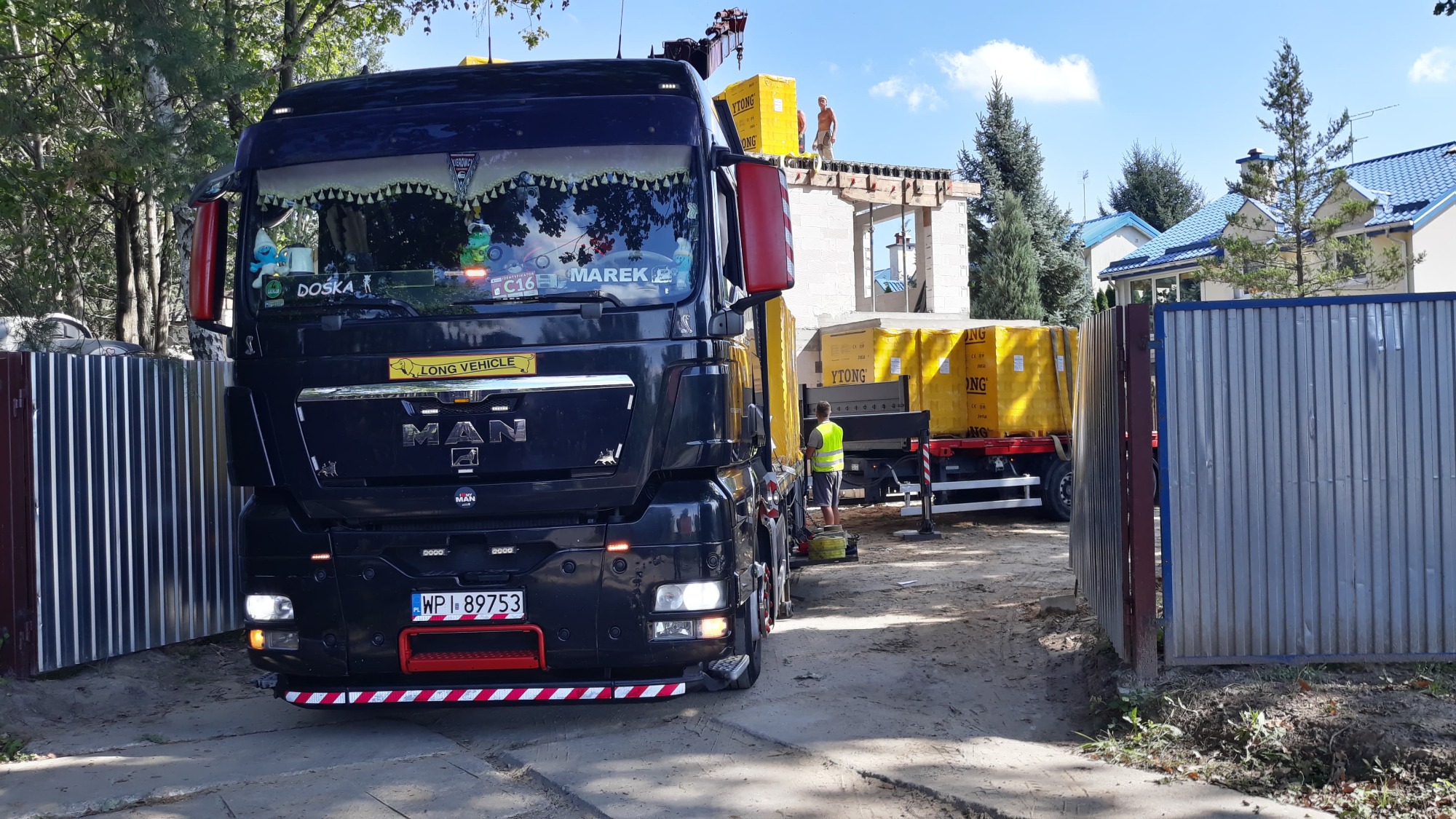Czarny ciągnik MAN z naczepą, z ładunkiem palet YTONG, dostarcza materiały budowlane na budowę domu jednorodzinnego; widoczni dwaj robotnicy na budowie. Na szybie ciągnika dekoracje i napisy...
