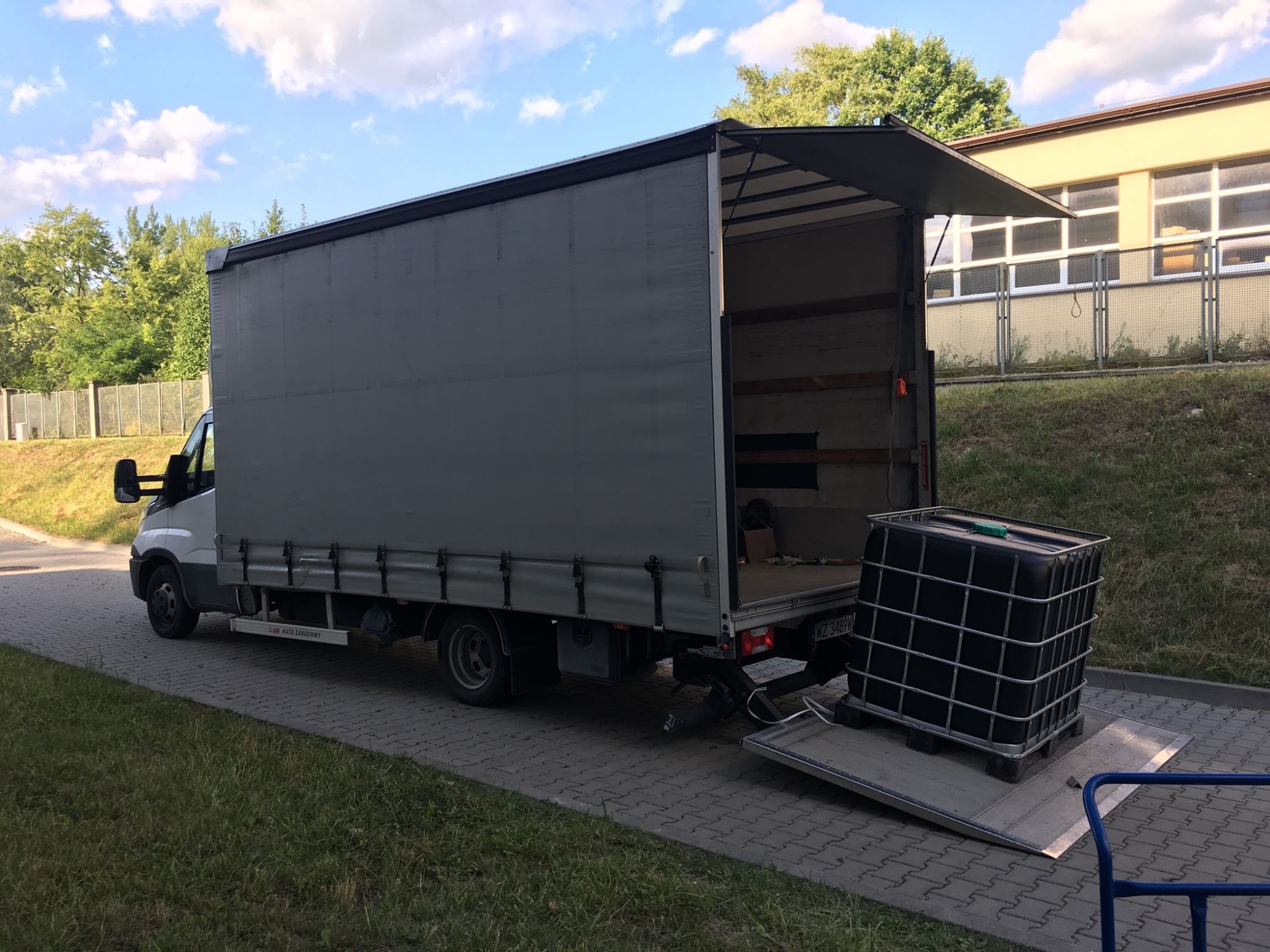 Szary samochód dostawczy z otwartą tylną klapą i rampą, na której znajduje się czarny pojemnik IBC na palecie, gotowy do załadunku.