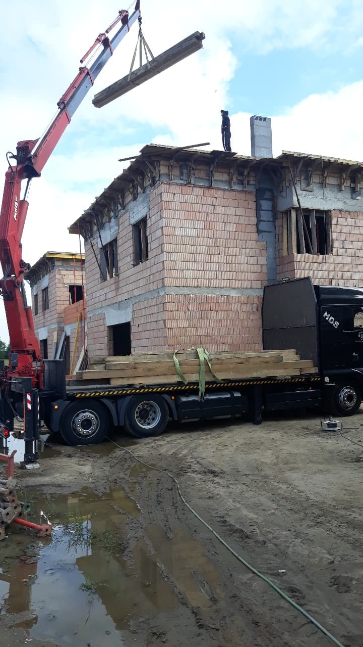 Ciężarówka z dźwigiem podnosi drewniane belki na budowę domu z cegły, pracownik na dachu, błotniste podłoże.