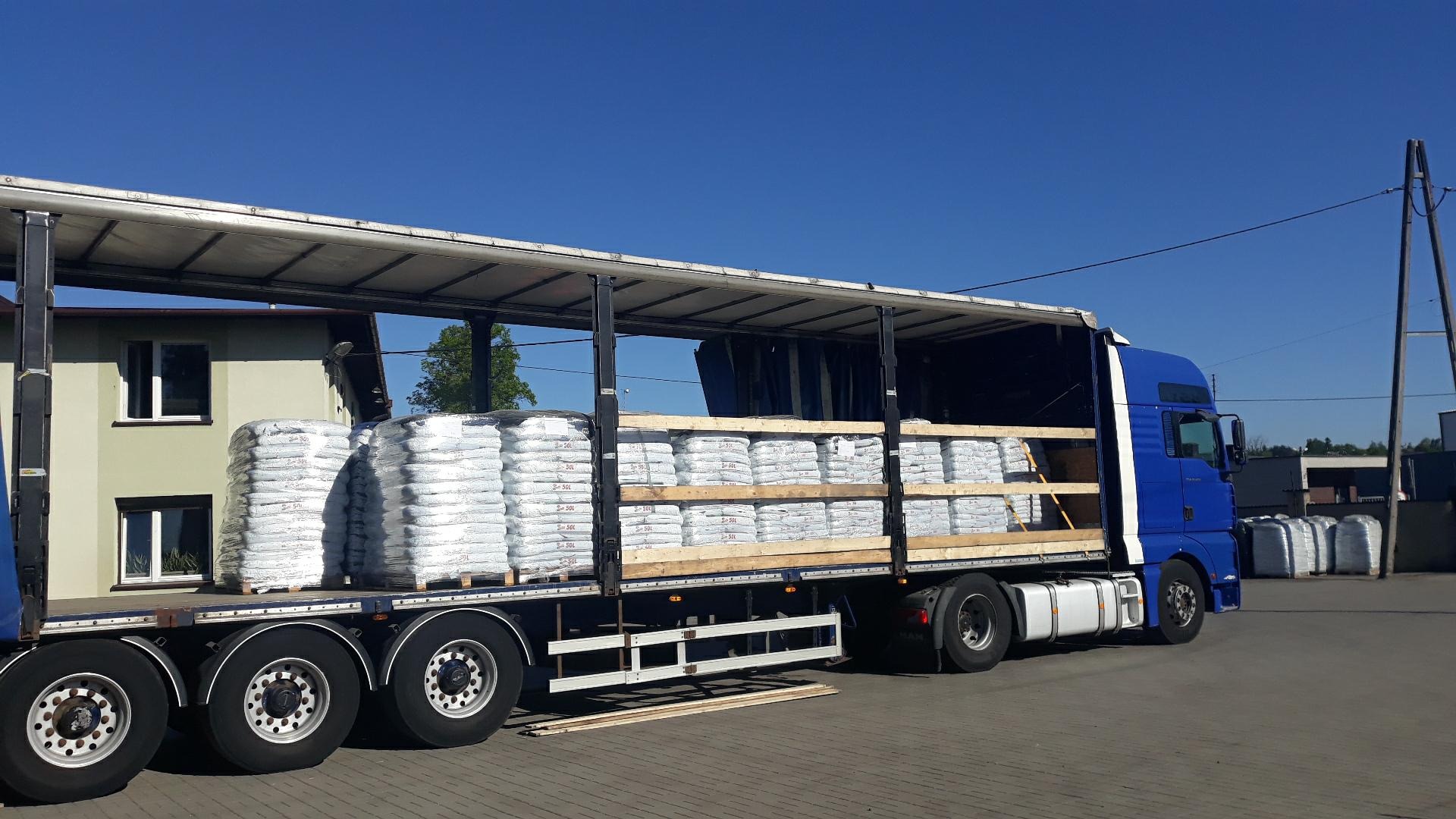 Ciężarówka z otwartą plandeką załadowana paletami z workami, widoczne zabezpieczenia ładunku z drewnianych belek, w tle budynek i słupy energetyczne.