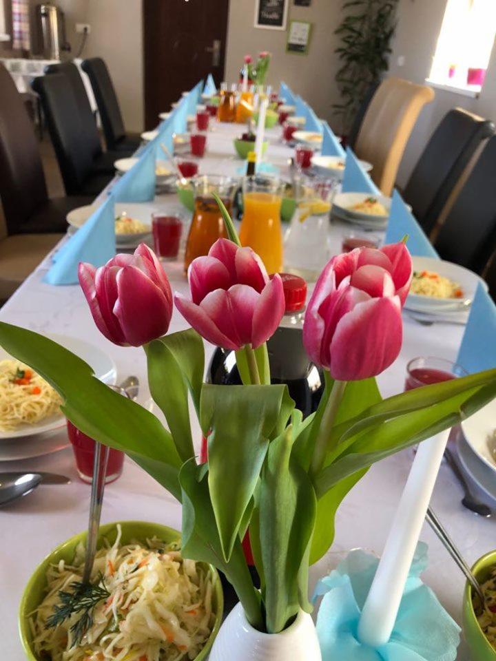 Ustawiony stół z białym obrusem, niebieskimi serwetkami, napojami w szklankach, sałatkami w zielonych miseczkach i talerzach z makaronem. W centrum wazon z czerwonymi tulipanami i butelką napoju.