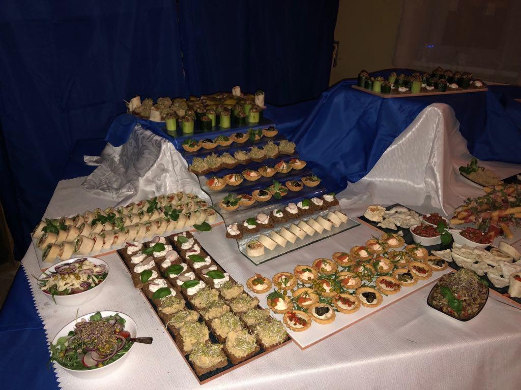 Bogato zastawiony stół cateringowy z różnorodnymi przekąskami: kanapeczki, tartaletki, sałatki, roladki, ułożone na białym obrusie z dekoracyjnymi fałdami, tło z granatowej tkaniny.