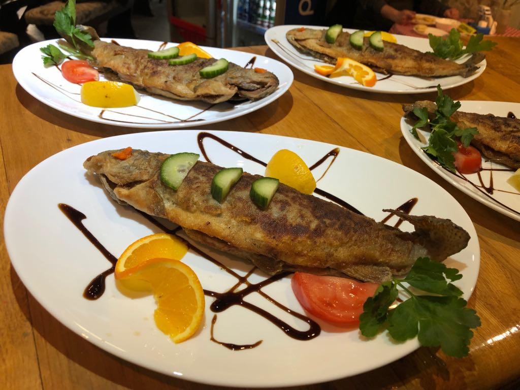 Smażona ryba, prawdopodobnie pstrąg, udekorowana plasterkami ogórka, pomarańczy, cytryny, pomidora i sosem balsamicznym, podana na białym talerzu, na drewnianym stole.