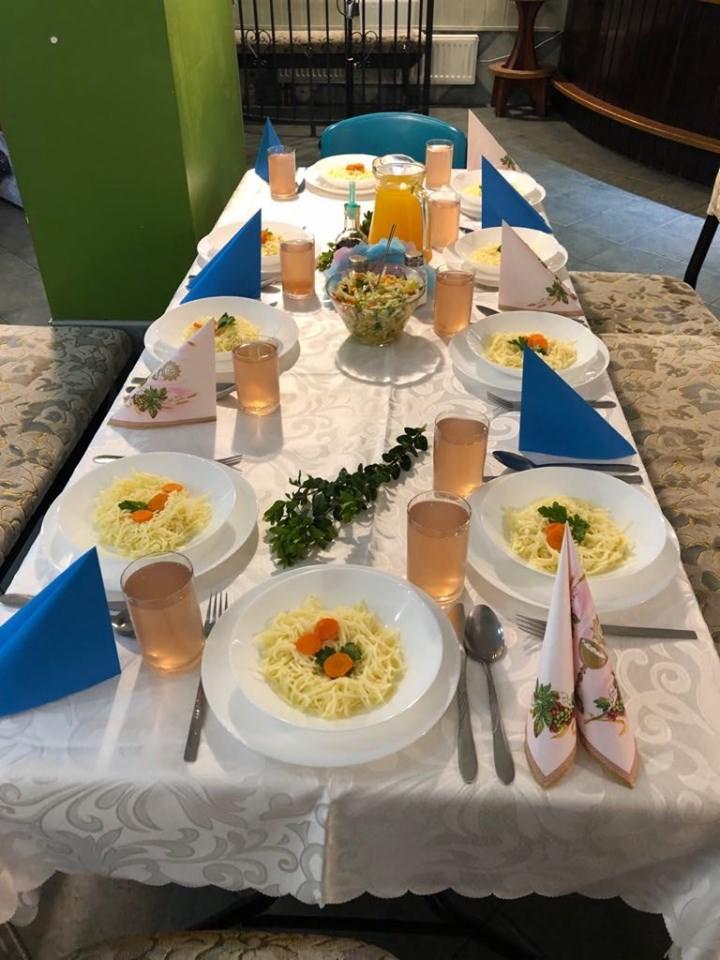 Ustawiony stół z białym obrusem, talerzami z makaronem z marchewką, szklankami z napojem, sałatką w szklanej misce i kolorowymi serwetkami.