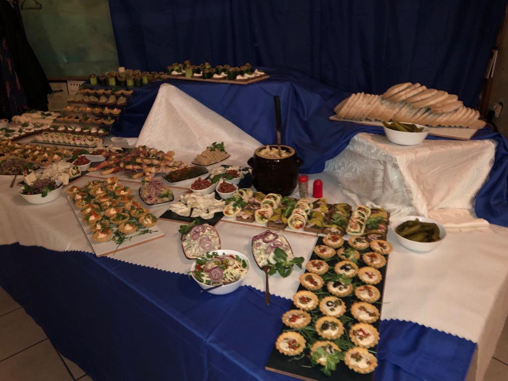 Stół z różnorodnymi przekąskami: tartaletki z oliwkami, roladki z warzywami, sałatki w miseczkach, kanapki i naczynie z gulaszem, ustawione na białym obrusie z niebieską falbaną.