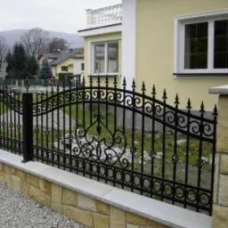 Czarne, ozdobne przęsło ogrodzenia z kutego żelaza, osadzone na niskim murku z kamienia naturalnego, na tle domu z żółtą elewacją i widokiem na zalesione wzgórza w oddali.