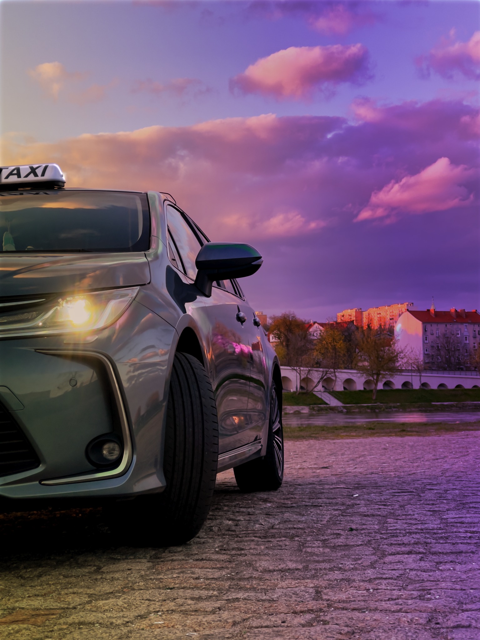 Szary samochód taxi z włączonymi światłami na tle fioletowo-różowego nieba i miejskiego krajobrazu z mostem.
