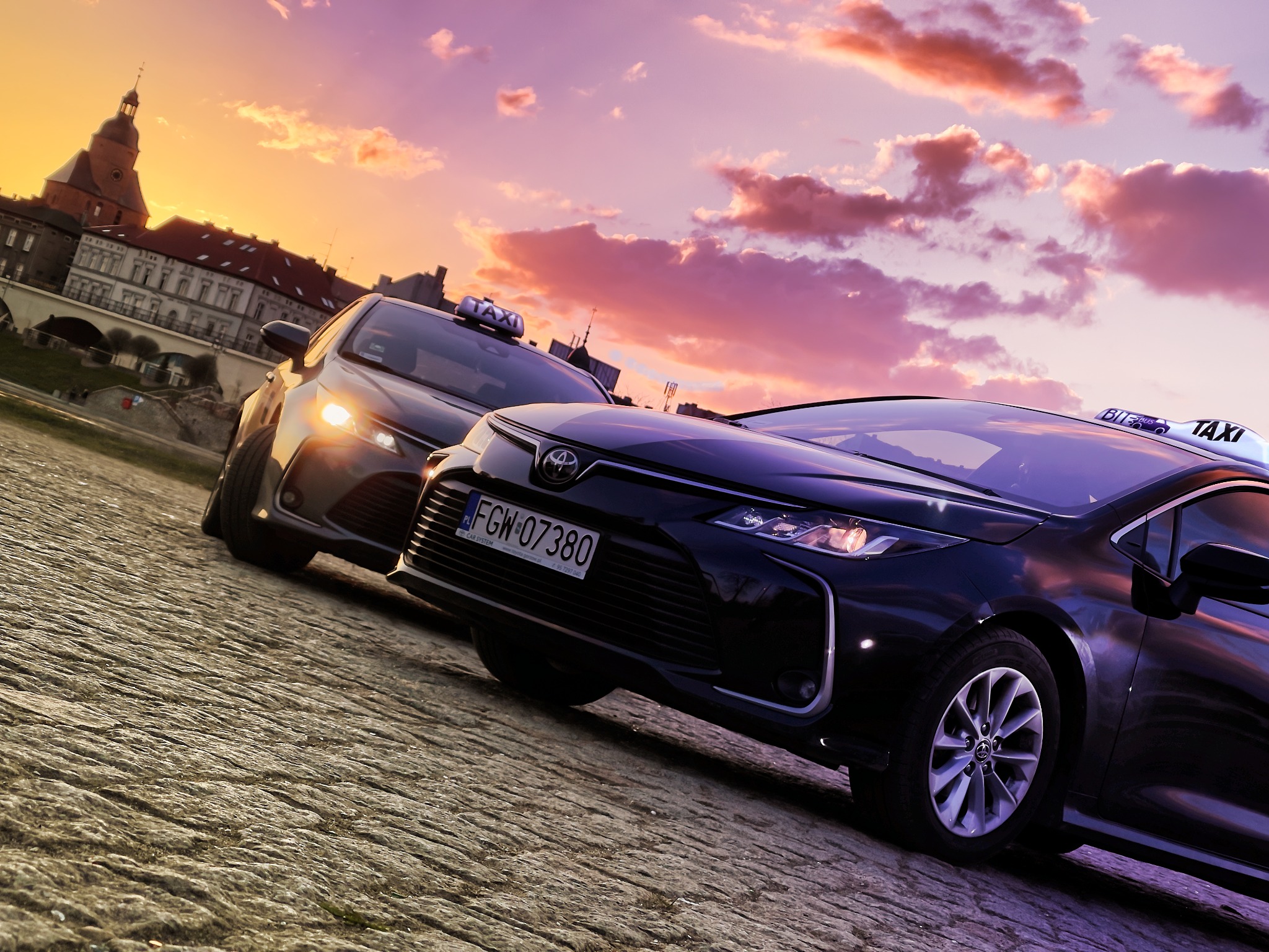 Dwa samochody taxi na tle zachodzącego słońca i zabytkowej architektury, widoczne oznaczenia taxi na dachach, tablice rejestracyjne z polskim numerem.