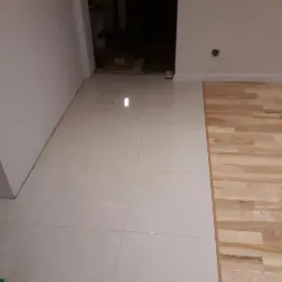 Połączenie błyszczących, kremowych płytek podłogowych i jasnego parkietu z naturalnego drewna w pomieszczeniu z białymi ścianami, widoczny fragment gniazdka elektrycznego.