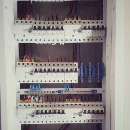 Rozdzielnia elektryczna z zamontowanymi bezpiecznikami i przewodami wewnątrz białej obudowy, widoczne różne kolory przewodów i oznaczenia na bezpiecznikach.