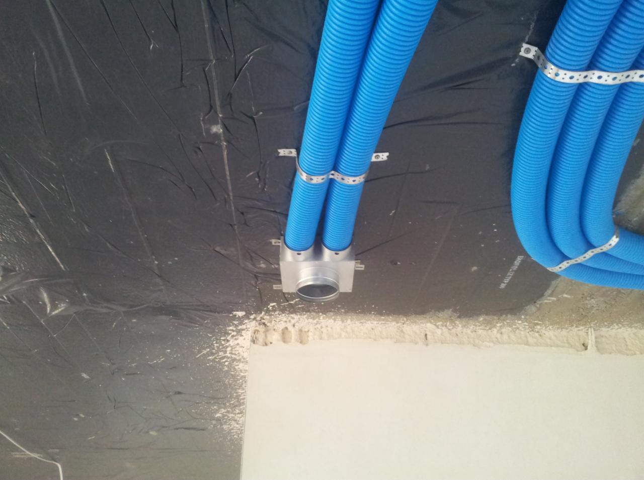 Niebieskie, karbowane rury wentylacyjne przymocowane do sufitu pokrytego czarną folią, widoczny fragment ściany w stanie surowym.