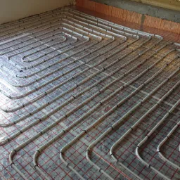 Białe rury ogrzewania podłogowego ułożone spiralnie na folii z czerwoną siatką, widoczne mocowania rur i surowy stan wykończenia podłoża.