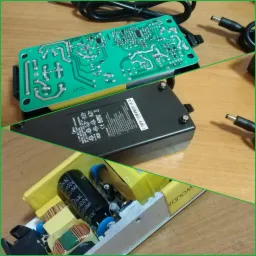 Otwarty zasilacz laptopa z widoczną płytką drukowaną, kondensatorami i elementami elektronicznymi, leżący na drewnianym blacie.