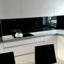 Nowoczesna kuchnia z białymi szafkami i czarnym panelem szklanym nad blatem, czajnik na płycie indukcyjnej, widok z jadalni z rośliną doniczkową na stole.