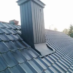 Nowy, ciemnoszary komin z blachy na dachu pokrytym ciemną dachówką karpiówką, widoczne elementy uszczelniające wokół komina.