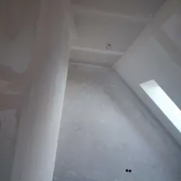 Poddasze w trakcie remontu: ściany po gładziowaniu, widoczne niedoskonałości i przygotowanie do malowania, okno dachowe w skosie, narzędzia pracy i materiały budowlane na podłodze.