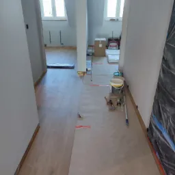 Korytarz w trakcie remontu: podłoga zabezpieczona papierem, widoczne narzędzia malarskie i puszki z farbą, ściany przygotowane do malowania, okna dachowe w tle.