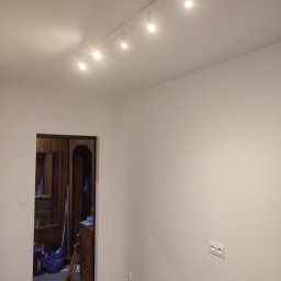 MIX MIR USŁUGI REMONTOWO WYKOŃCZENIOWE MIROSŁAW KALINKA - Białe ściany po malowaniu z widoczną listwą przypodłogową i oświetleniem sufitowym na szynie, w tle widoczne otwarte drzwi do zagraconego pokoju z boazerią.