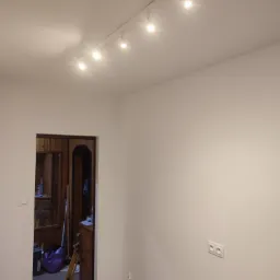 Białe ściany po malowaniu z widoczną listwą przypodłogową i oświetleniem sufitowym na szynie, w tle widoczne otwarte drzwi do zagraconego pokoju z boazerią.