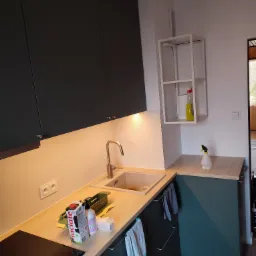 Nowoczesna kuchnia z ciemnozielonymi szafkami, drewnianym blatem, stalową baterią i widocznymi produktami spożywczymi na blacie. Oświetlenie podszafkowe dodaje ciepła, a biała półka na ścianie...