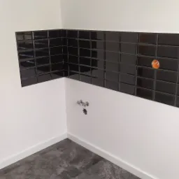 Fragment łazienki po remoncie: ciemne, błyszczące płytki ułożone na ścianie nad białym pasem, szara podłoga z płytek imitujących kamień, widoczne przyłącza wodne.