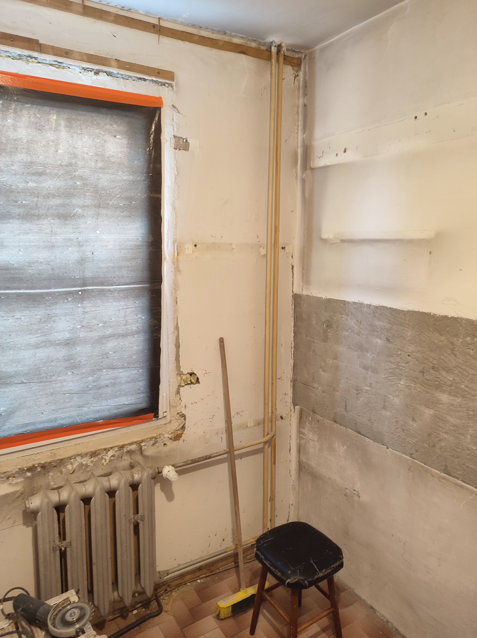 Pokój w trakcie prac przygotowawczych do remontu: zabezpieczone okno folią, odsłonięta część ściany, stara instalacja grzewcza z grzejnikiem żeliwnym, widoczne narzędzia i drabina.