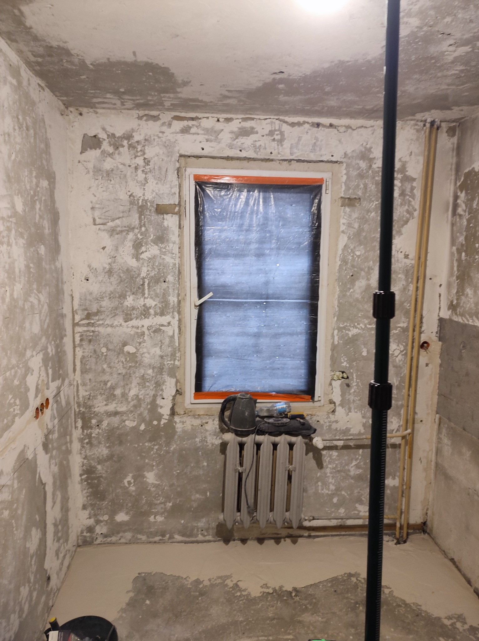 Pokój w trakcie przygotowania do prac wykończeniowych: ściany z widocznymi śladami po starych powłokach, okno zabezpieczone folią, grzejnik pod oknem, pionowy słup podpierający.