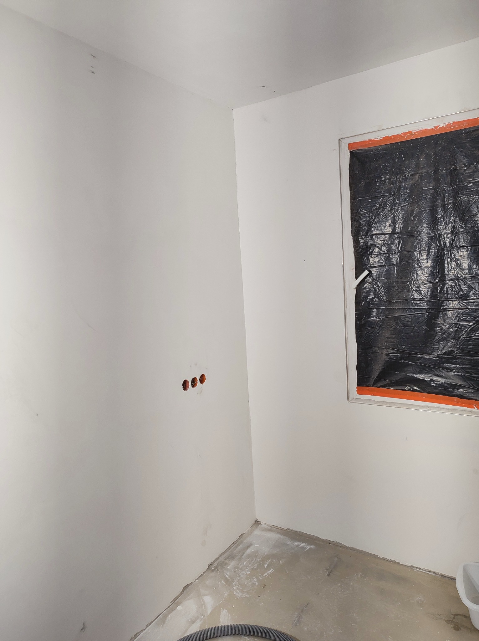 Pokój w trakcie przygotowań do malowania: okno zabezpieczone folią, ściany zagruntowane, widoczne puszki instalacyjne elektryczne.